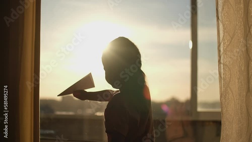 A child plays with a paper plane near the window of the house during sunset.Authentic scene.