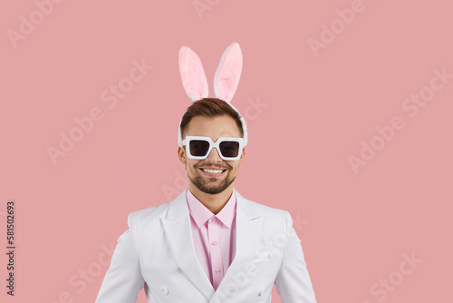 Young happy Caucasian man with funny bunny rabbit ears on head and sunglasses smiling broadly looking at camera dressed in classic white suit stands on pink background. Easter concept © Studio Romantic