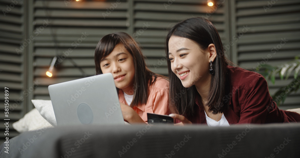 Two asian sisters lying on bed with laptop, having online shopping, cheerfully smiling and chatting, online shopping, girls spending time together, having online shopping. family time, online shopping