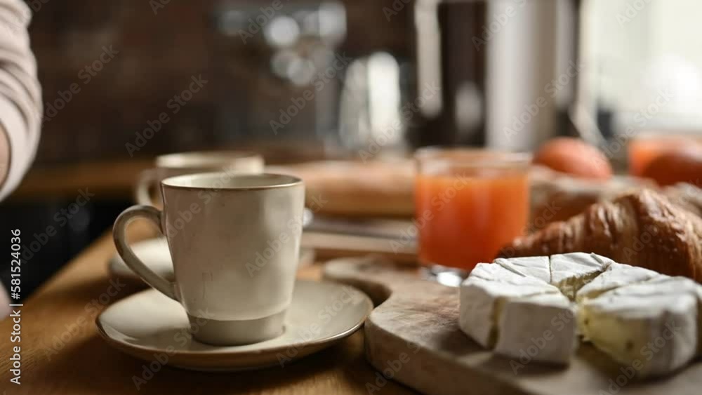 Girl hand put coffee to cups for breakfast with croissants and brie cheese at sunny kitchen. Morning meal preparation with cappuccino espresso beverage in mugs Girl hand put coffee to cups for breakfast with croissants and brie cheese at sunny kitchen. Morning meal preparation with cappuccino espresso beverage in mugs