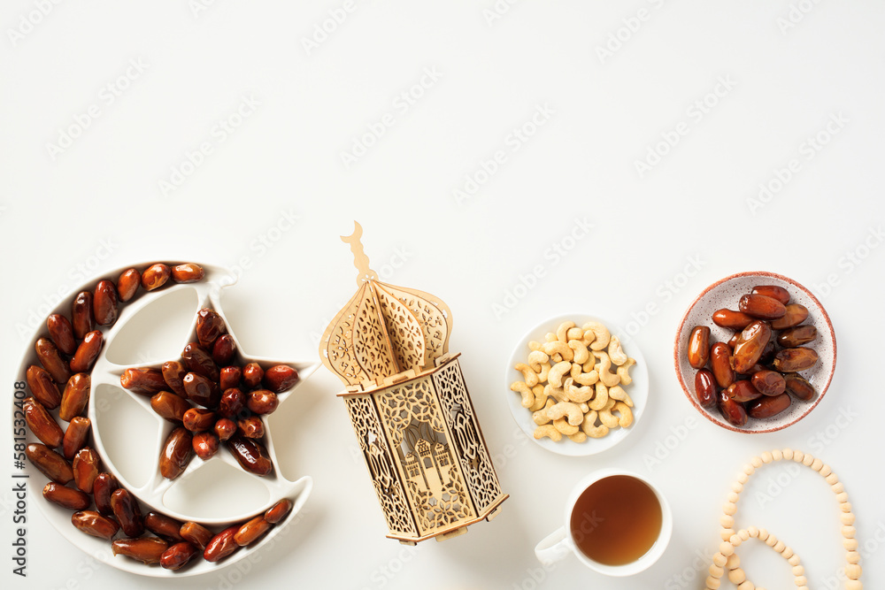Naklejka premium Muslim holiday Ramadan Kareem concept. Flat lay dried dates in Islamic star and crescent plate, nuts, oriental lantern, cup of tea, rosary on white background.