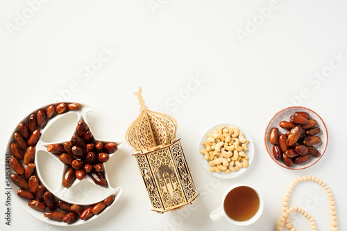 Muslim holiday Ramadan Kareem concept. Flat lay dried dates in Islamic star and crescent plate, nuts, oriental lantern, cup of tea, rosary on white background.