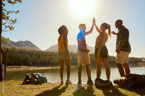 Rear View Group Of Friends With Backpacks On Vacation Hiking By Lake And Mountains Giving High Five