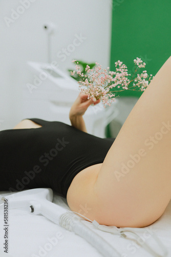 Beauty master makes laser hair removal of legs close-up for a beautiful girl in a black bodysuit with flowers in her hands in a beauty salon