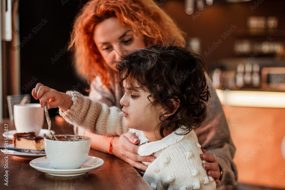 Beautiful young red-haired mother with cute curly-haired daughter are sitting in cozy cafe and drinking hot school. Mothers Day. Warm.