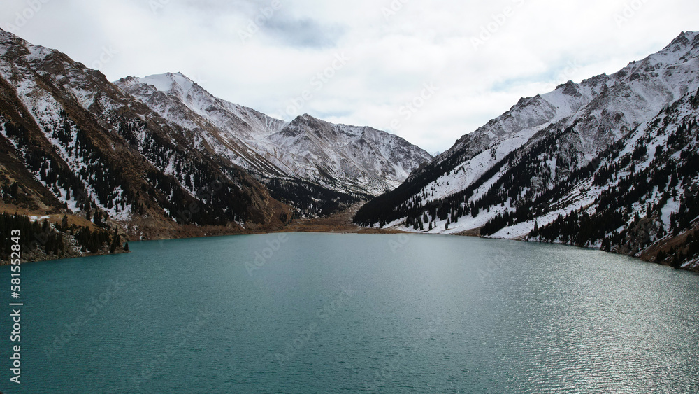 Obraz premium A lake in the mountains with turquoise blue water. Drone view of clear water, coniferous trees and snowy mountains. People walk along the shore, low bushes grow. Big Almaty lake. Kazakhstan