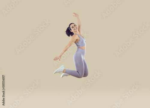 Happy athletic woman jumping in fashionable lilac sportswear isolated on beige background. Full length portrait of cheerful excited young sportswoman with perfect figure having fun jumping high.