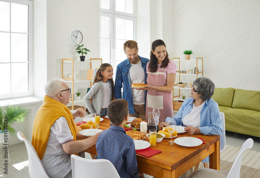 Dinner in big friendly happy family with parents, kids and grandparents at table in living room. Young mom holding a pie and looking at grandma with smile. Emotional family moments together concept. Dinner in big friendly happy family with parents, kids and grandparents at table in living room. Young mom holding a pie and looking at grandma with smile. Emotional family moments together concept.