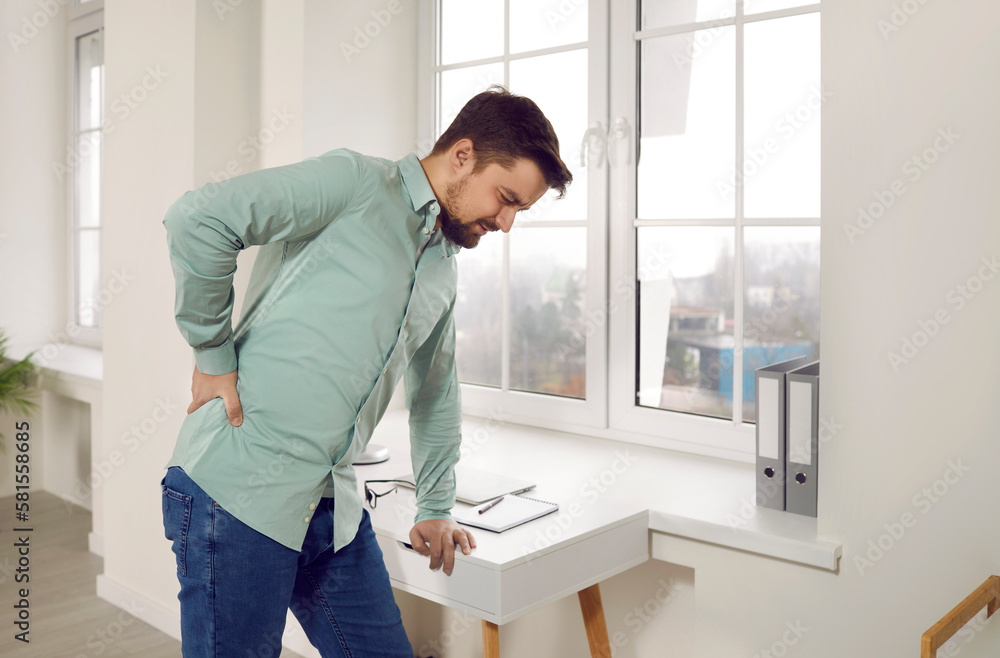 Handsome oung man suffering from backache in ofiice. Office worker standing leaning table touching his painful lower back, man feeling pain or spinal spasm. Sedentary life, office syndrome concept