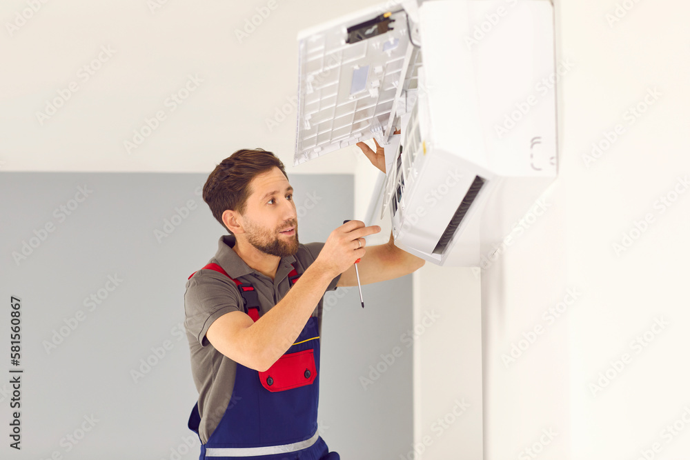 Male worker installing or fixing parts in air conditioner. Young man in uniform standing on ladder and installing new white air conditioner on wall. AC installation and maintenance service concept Male worker installing or fixing parts in air conditioner. Young man in uniform standing on ladder and installing new white air conditioner on wall. AC installation and maintenance service concept