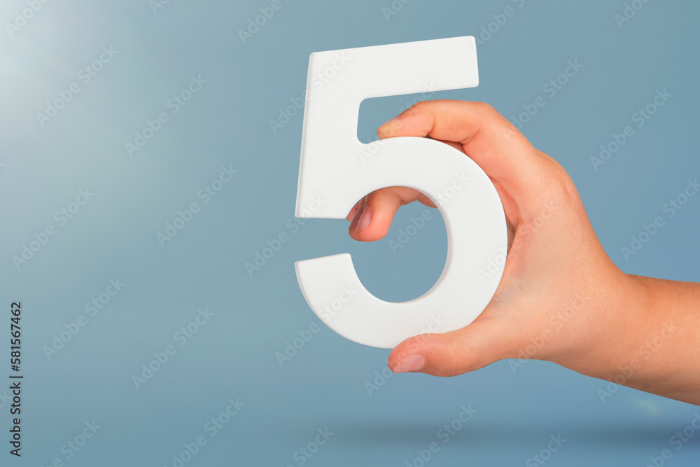 Number five in hand. Hand holding white number 5 on blue background with copy space. Concept with number five. Birthday 5 years, fifth grade, five day work week - obrazy, fototapety, plakaty 