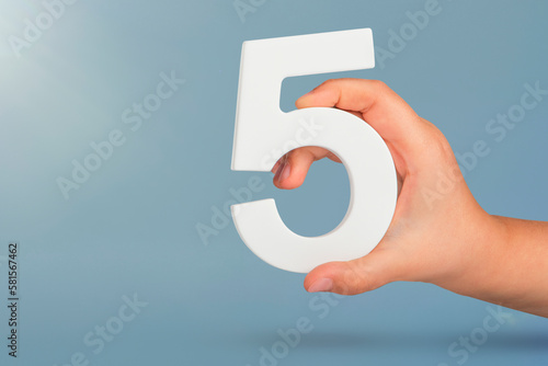 Number five in hand. Hand holding white number 5 on blue background with copy space. Concept with number five. Birthday 5 years, fifth grade, five day work week photo