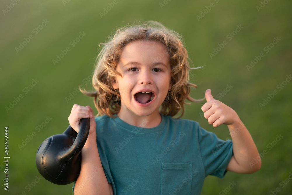 Child lifting the kettlebell in park outside. Sport activities at leisure with children. Sporty kid boy holding dumbbells kettlebell. Child lifting the kettlebell in park outside. Sport activities at leisure with children. Sporty kid boy holding dumbbells kettlebell.