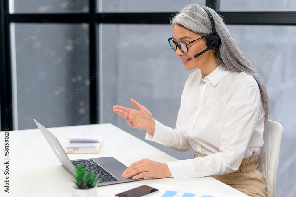 Mature senior office worker businesswoman with a pleasant smile in headset and glasses looking at laptop screen, sitting at the desk. Female call center operator is feeling motivated and ready to work