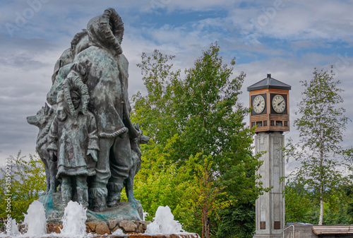 Fairbanks, Alaska, USA - July 27, 2011: Unknown First Family statue and fountain on Golden Heart Plaza. Child and father side, with Rotary Clock Tower and green foliage under blue cloudscape photo