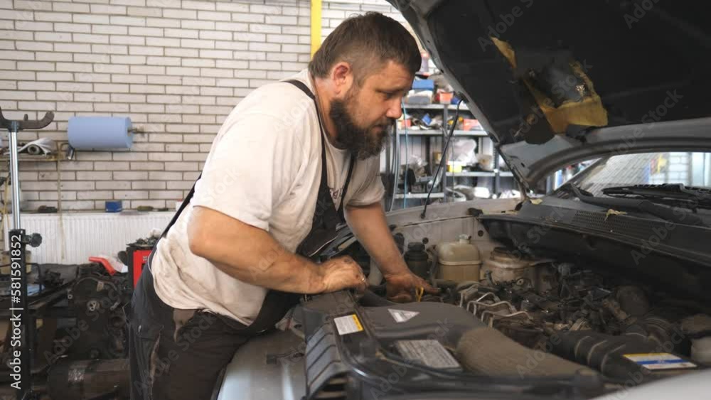 Professional auto mechanic works under hood of car. Repairman fixing engine of a vehicle at workshop or service. Bearded man checking automobile motor at garage. Concept of maintenance and diagnostic Professional auto mechanic works under hood of car. Repairman fixing engine of a vehicle at workshop or service. Bearded man checking automobile motor at garage. Concept of maintenance and diagnostic