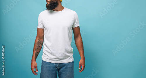 Portrait of handsome, young, stunning, perfect bearded guy wearing a white blank t-shirt stands against turquoise background. T-shirt template.