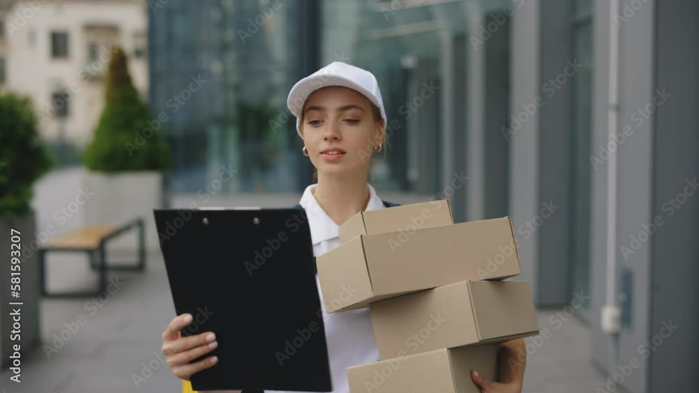 Caucasian Female Courier Going Near the Busines Office, Carries Packages and a Tablet Folder with the Addresses of Customers. Delivery Worker on the Way to Deliver Postal Parcel to a Client Caucasian Female Courier Going Near the Busines Office, Carries Packages and a Tablet Folder with the Addresses of Customers. Delivery Worker on the Way to Deliver Postal Parcel to a Client