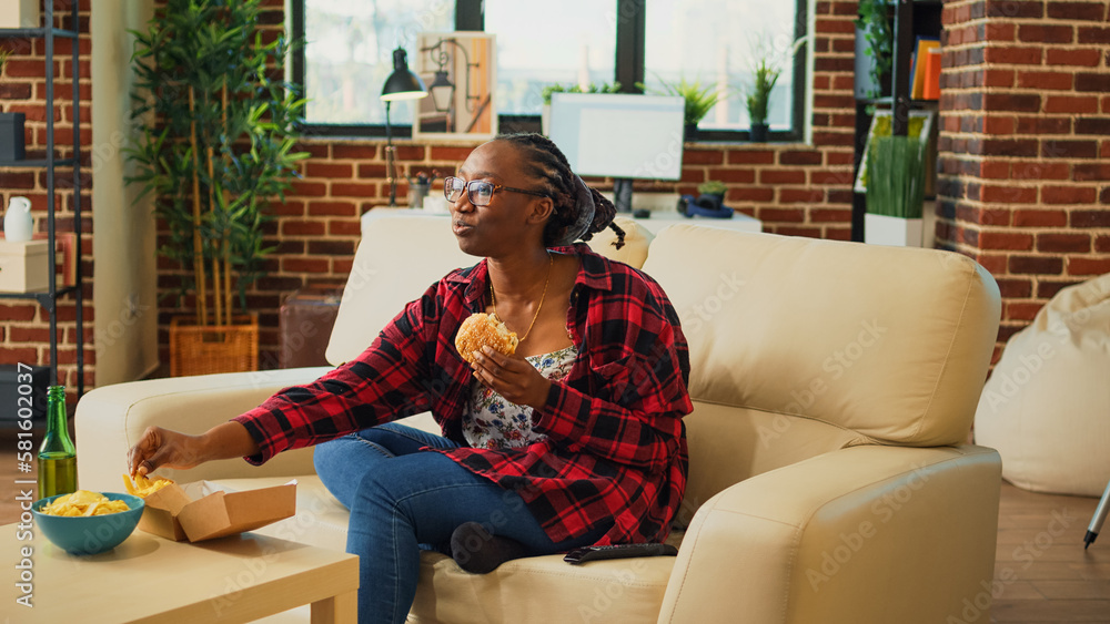 African american person taking bite of burger on couch, eating fast food from delivery a home. Young adult enjoying takeaway meal with fries and hamburger, watch movie on television.