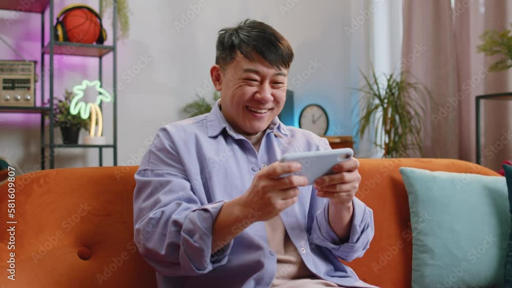 Worried asian handsome man enthusiastically playing racing or shooter video games on smartphone at home. Chinese guy using smartphone gadget app with drive simulator sitting on sofa in living room Worried asian handsome man enthusiastically playing racing or shooter video games on smartphone at home. Chinese guy using smartphone gadget app with drive simulator sitting on sofa in living room