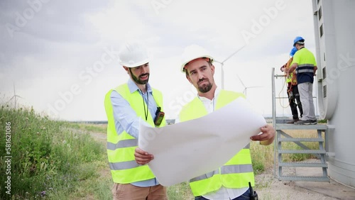 Two windmill engineers looking a blueprint with the plan of the construction of the electricy turbine. At background workers or contractors doing their job . Renewable energy concept. Slow motion photo
