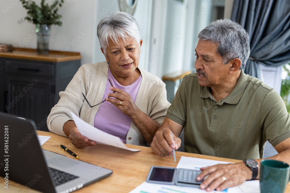 Biracial senior couple with calculator and bills analyzing finances and expenses on table at home Biracial senior couple with calculator and bills analyzing finances and expenses on table at home