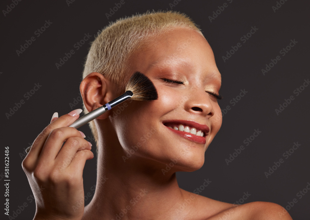 Obraz premium Woman, smile and makeup brush for beauty cosmetics, skincare or facial treatment against a gray studio background. Happy beautiful female model applying cosmetic product on face for self care or love