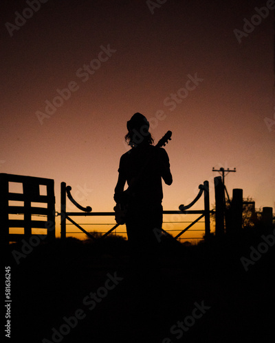 Foto artística Contraluz de hombre tocando la guitarra bajo un cielo rojo y naranja delante de una tranquera durante el atardecer de la llanura pampeana musico guitarrista pampeano photo