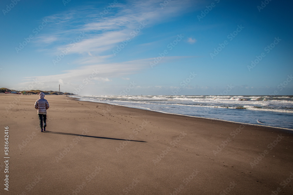 mujer con campera caminando en la playa al costado con luz de dia en invierno con campera por la costa