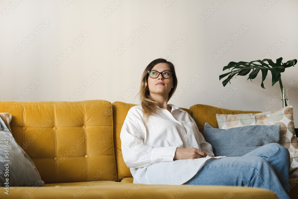 Portrait of young caucasian woman with long hair wearing glasses, casual clothing sitting on yellow sofa at home in modern interior looking away. Copy space. Portrait of young caucasian woman with long hair wearing glasses, casual clothing sitting on yellow sofa at home in modern interior looking away. Copy space.