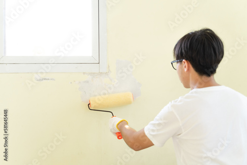 Asian young adult man using roller painting for painting wall to interior renovation wall house. Painter man painting wall by himself at home. Worker repair home decoration and renovation concept.