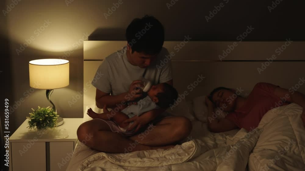 stressed father trying to feeding milk bottle to his crying newborn baby while his wife sleeping on a bed at night stressed father trying to feeding milk bottle to his crying newborn baby while his wife sleeping on a bed at night