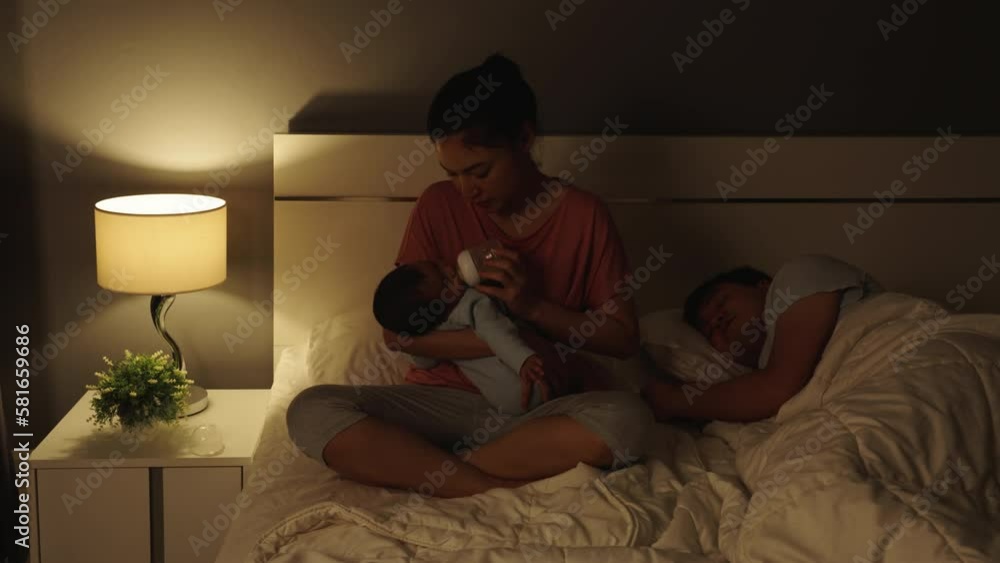 mother feeding milk bottle to newborn baby while her husband sleeping on a bed at night mother feeding milk bottle to newborn baby while her husband sleeping on a bed at night