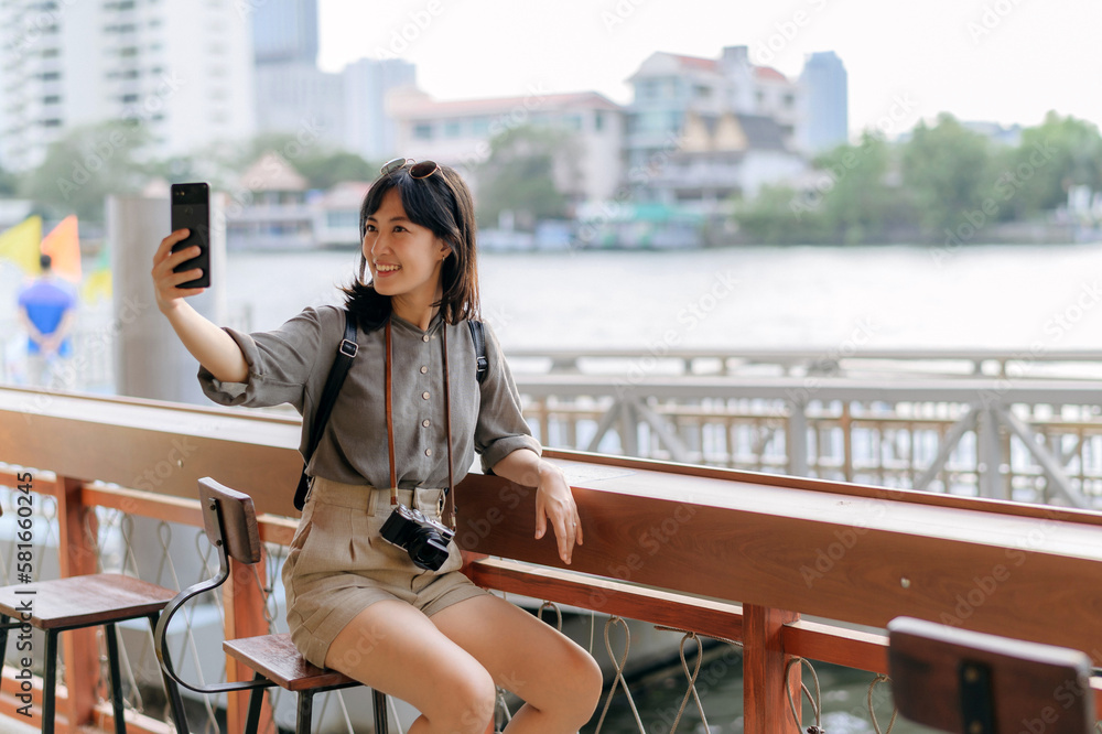 Young Asian woman backpack traveler using mobile phone in express boat pier on Chao Phraya River in Bangkok. - obrazy, fototapety, plakaty 