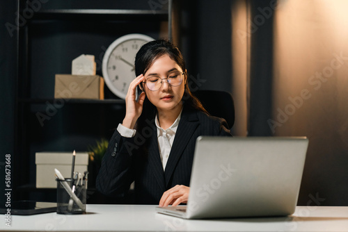 Stressed asian business woman working late at night in the office hands on head feeling headache. Tired woman looking at laptop screen working hard sitting in the dark room office. Overtime concept