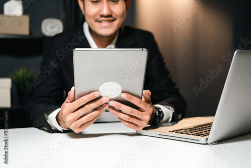 Young asian businessman working with laptop computer in the dark office at night. Attractive handsome Indian man work hard overtime in home with floor lamp ambient warm light late at night