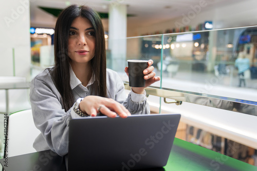 Start of the working day with coffee in the cafe. A young woman drinks coffee and works at a laptop in a shopping center cafe