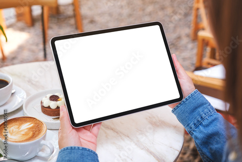 Mockup image of a woman holding digital tablet with blank white desktop screen with cake and coffee cup on the table in cafe