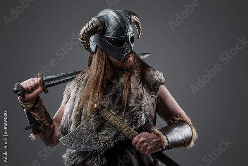 Portrait of nordic barbarian dressed in fur and horned helmet holding sword and axe.