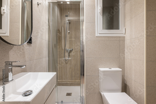 Modern small bathroom with stylish beige tiles  glass shower cabin  sink  toilet and window. Concept of multifunctional bathroom in hotel. Young family apartment