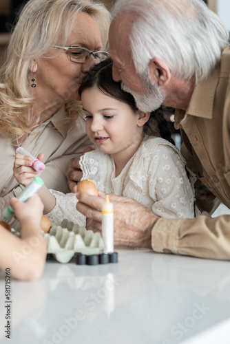 Grandparents kissing their granddaughters head while she decorates Easter eggs with her brother