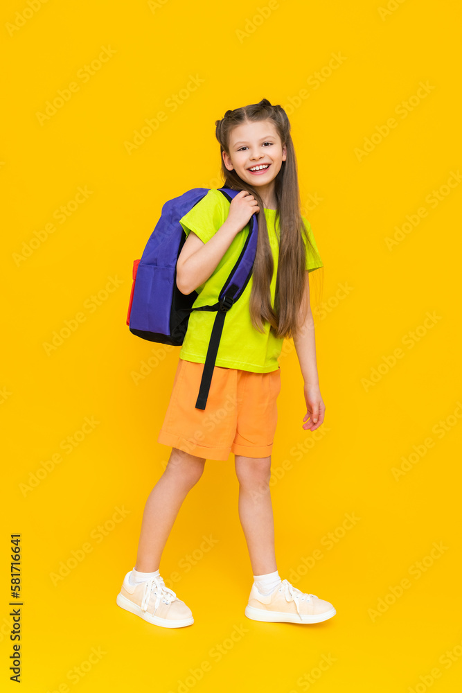 A child with a full-length backpack. A little girl with a satchel in shorts. Additional educational summer classes for children. Preparation for the school year. Yellow isolated background. A child with a full-length backpack. A little girl with a satchel in shorts. Additional educational summer classes for children. Preparation for the school year. Yellow isolated background.