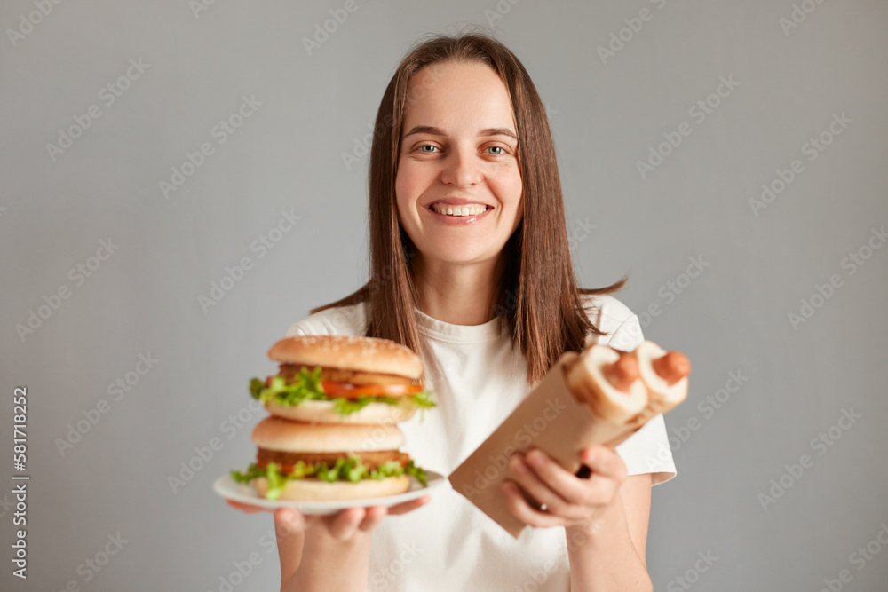 Horizontal shot of smiling satisfied female with brown hair, standing isolated on gray background, holding hot dogs and american hamburger, enjoying fast food.