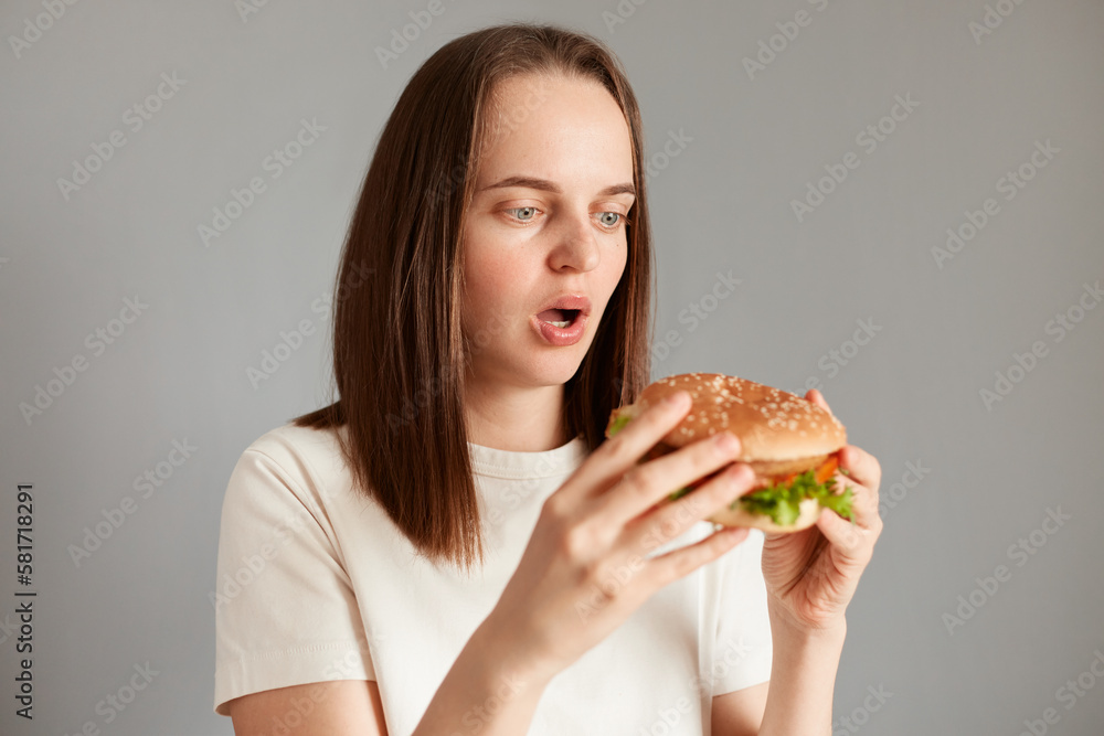 Proper nutrition, healthy fast food, unhealthy choice. Young amazed astonished woman wearing white t-shirt holding in hand burger isolated on gray background.