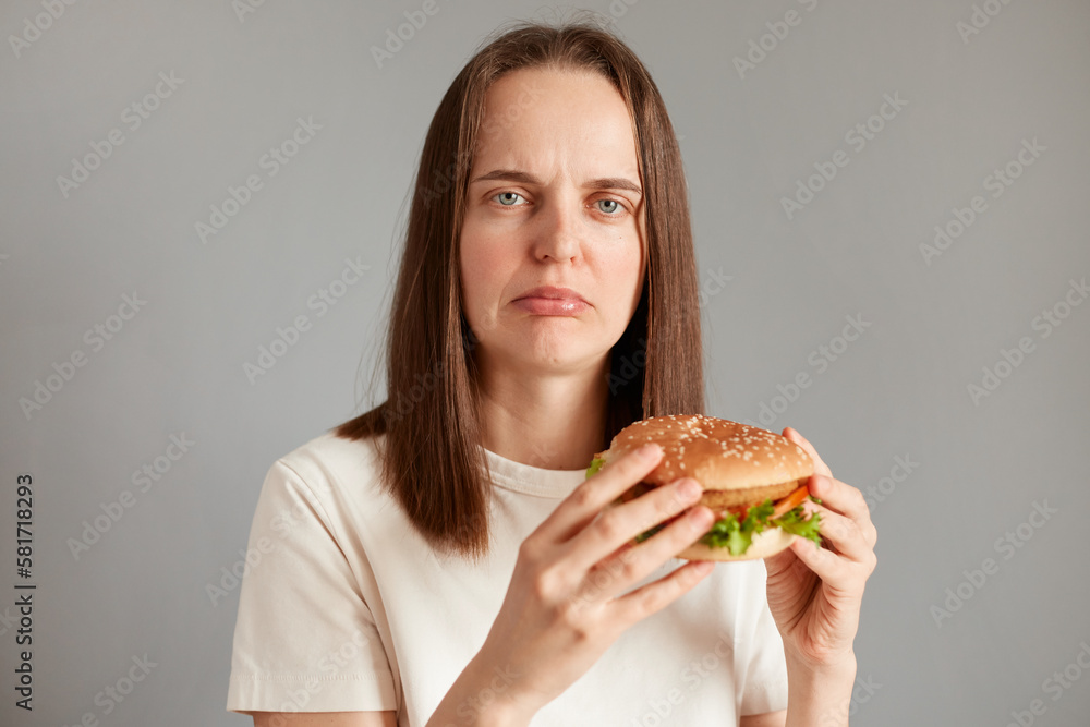 Indoor shot of sad attractive woman keeps diet, feels hungry, holding american hamburger in hands, looking at camera with sorrow, needs to keep healthy eating.