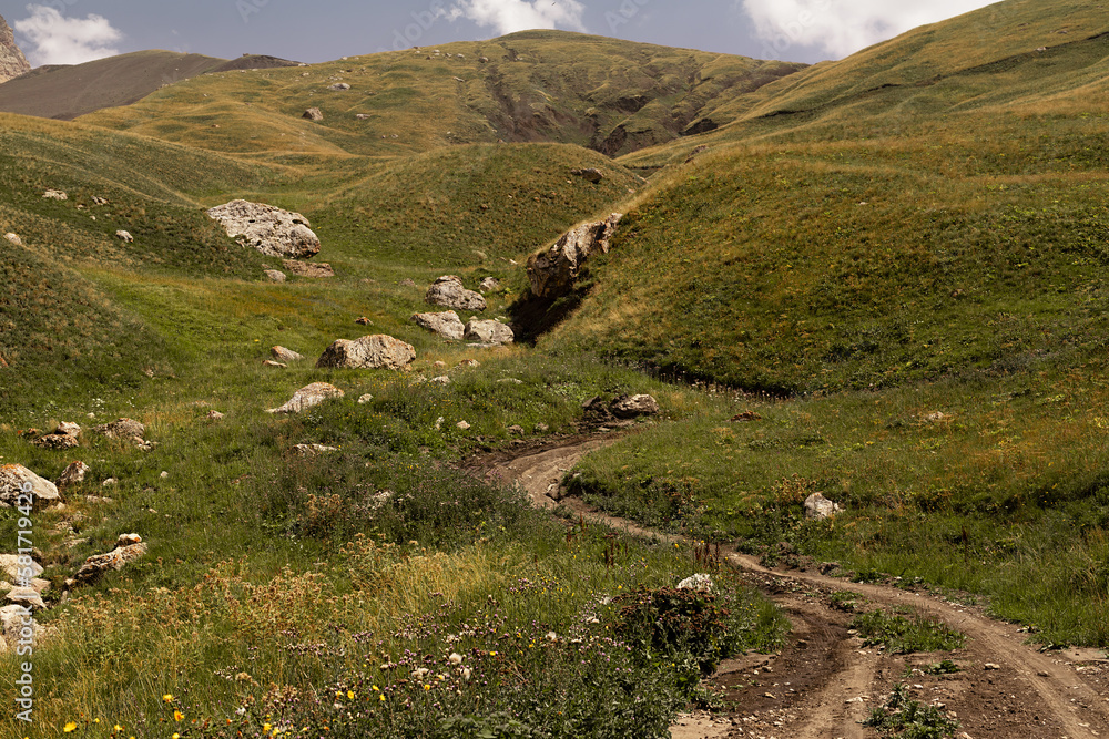 Obraz premium Mountain landscape - hilly mountain valley with country road and big boulders in fine sunny summer day, green and yellow velvety meadow on with slopes. Awe travel in Dagestan.