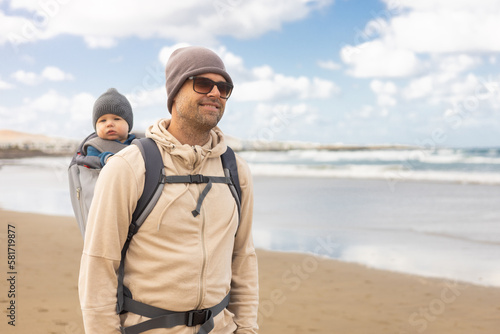 Young father carrying his infant baby boy son in backpack on windy sandy Famara beach, Lanzarote island, Spain. Family travel and winter vacation concept © kasto