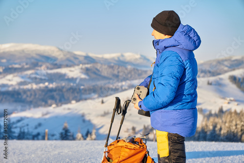 Tourist travelling in winter dressed in winter sportswear with backpack walking with trekking poles in the snowy pine mountains. Traveller with his backpack looking on the hill. 