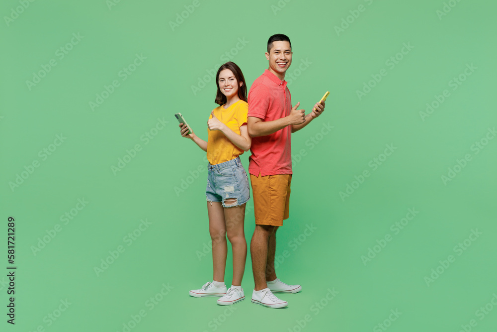 Full body young couple two friends family man woman wear basic t-shirts together hold in hand use mobile cell phone stand back to back isolated on pastel plain light green background studio portrait. Full body young couple two friends family man woman wear basic t-shirts together hold in hand use mobile cell phone stand back to back isolated on pastel plain light green background studio portrait.