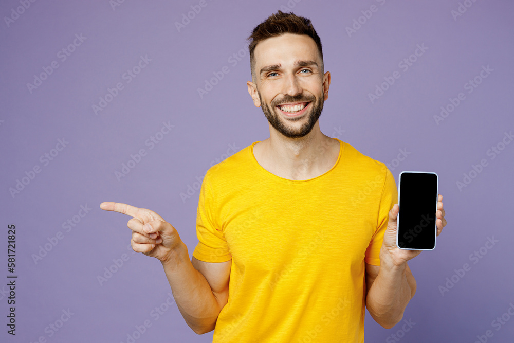 Young man wear yellow t-shirt hold in hand use mobile cell phone with blank screen workspace area point finger aside isolated on plain pastel light purple background studio portrait Lifestyle concept Young man wear yellow t-shirt hold in hand use mobile cell phone with blank screen workspace area point finger aside isolated on plain pastel light purple background studio portrait Lifestyle concept